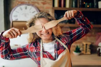 Playful housewife in an apron covers her eyes with wooden spatulas, kitchen interior on background. Happy female cook making healthy vegetarian food, salad cooking