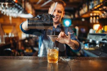 Male bartender in apron pours a drink through a sieve into a glass. Barman at the bar counter. Alcohol beverage preparation