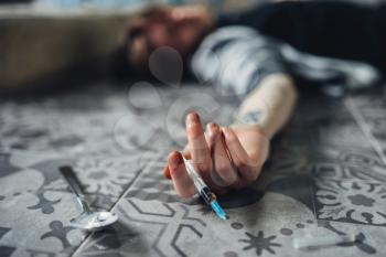 Drug addict lies on the floor, hand with syringe and spoon for dose preparing on the table. Addiction concept, addicted people
