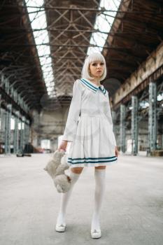 Cute anime style blonde woman with toy bear in hand. Cosplay, japanese culture, doll in dress on abandoned factory