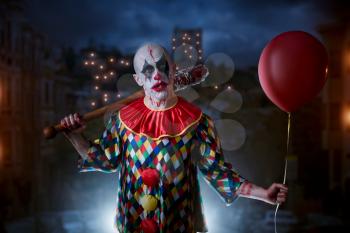 Scary bloody clown with baseball bat and air balloon, night city on background, nightmare. Man with makeup in carnival costume, crazy maniac