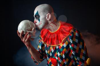Mad bloody clown holds human skull, horror. Man with makeup in carnival costume, crazy maniac