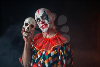 Bloody clown with crazy face holds human skull, terrible secret. Man with makeup in carnival costume, mad maniac