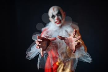 Mad bloody clown reaching for the victim with his hands, front view. Man with makeup in carnival costume, crazy maniac