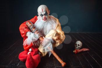 Scary bloody clown with baseball bat sitting on the floor, around the scattered parts of the human body. Man with makeup in carnival costume, crazy maniac
