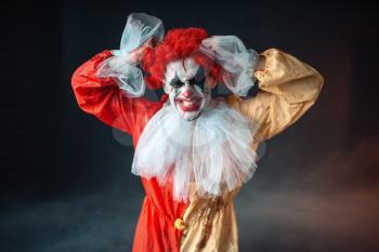 Scary bloody clown tears his hair, jerk in anger. Man with makeup in carnival costume, crazy maniac