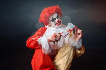 Scary bloody clown with crazy eyes looking on his knife. Man with makeup in carnival costume, mad maniac