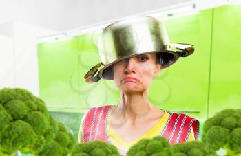 Crazy housewife in apron with a pot on her head among the broccoli, kitchen interior on background. Funny female person cooking