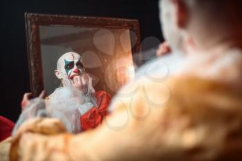 Frightened bloody clown with crazy eyes looking at the mirror, nightmare. Man with makeup in carnival costume, mad maniac