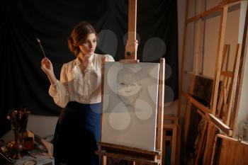 Cute female painter with brush standing against easel in studio. Creative paint, woman drawing pencil sketch, workshop interior on background