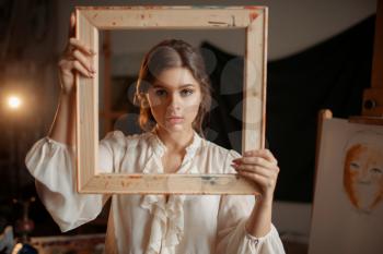 Female painter holds wooden frame at her face in studio, portrait drawing concept. Artist painting in class, workshop interior on background
