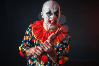 Mad bloody clown holds human hand, finger in his teeth. Man with makeup in halloween costume, crazy maniac 