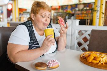 Fat woman eating doughnuts in fastfood restaurant, unhealthy food. Overweight female person at the table with junk dinner, obesity problem