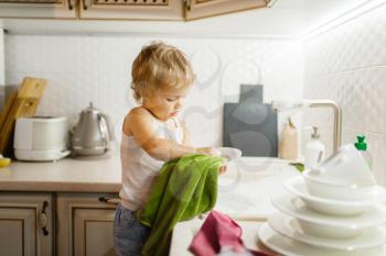 A little girl wipes the dishes on the kitchen. Baby doing housework at home. Young housewife clean the house