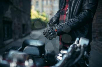 Male biker in leather jacket puts on gloves before riding on classical chopper, safety equipment