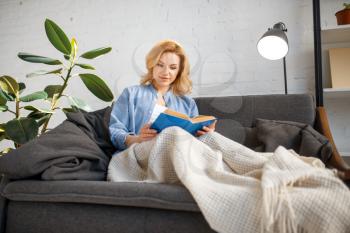 Young woman under a blanket reading a book on cozy yellow couch, living room in white tones on background. Attractive female person with magazine sitting on sofa at home