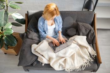 Young woman under a blanket using laptop on cozy yellow couch, living room in white tones on background. Attractive female person with magazine sitting on sofa at home