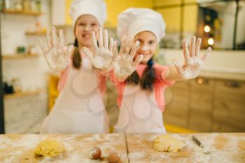 Two little girls cooks in caps shows hands covered in flour, cookies preparation on the kitchen. Kids cooking pastry, children chefs preparing cake
