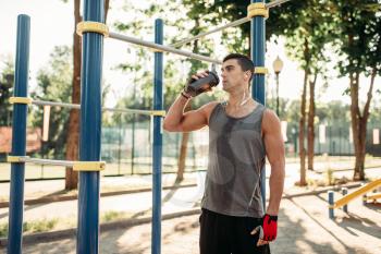 Male athlete in headphones drinks water after outdoor fitness training. Strong sportsman on sport workout in summer park