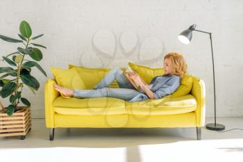 Young woman lying on cozy yellow couch and reading a book, living room in white tones on background. Attractive female person with magazine sitting on sofa at home