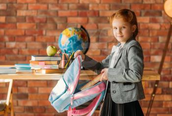 Cute schoolgirl puts textbook into the schoolbag, side view. Female pupil with backpack, desk with textbooks, apple and globe on backgrpund, young girl in the school