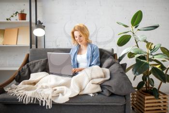 Young woman under a blanket using laptop on cozy yellow couch, living room in white tones on background. Attractive female person with magazine sitting on sofa at home