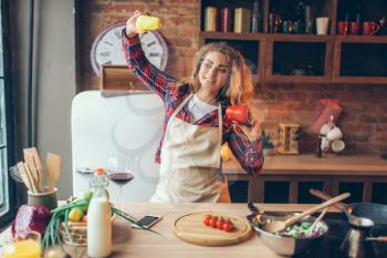 Smiling housewife in headphones and apron holding fresh pepper in hands and dance on the kitchen. Female kitchener cooking healthy vegetarian food, salad preparation