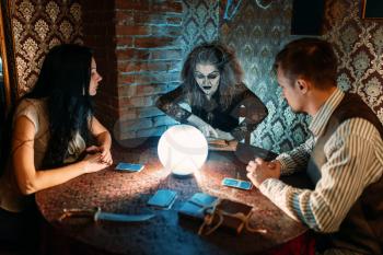 Young family and witch at the table with crystal ball on spiritual seance, scary wizard reads the spell. Female foreteller calls the spirits