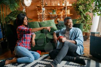 Black couple sitting on the floor and drinks coffee against a couch at home. Happy african love couple leisures in their house, cheerful family relaxing together in the morning