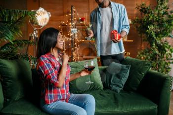 Black man with red rose and gift behind his woman with glass of wine on sofa, romantic meeting at home. Happy african love couple leisures in their house