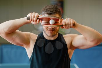 Man holds rackets and ping pong ball at his face, workout indoors. Male person in sportswear standing at the table with net, training in table-tennis club