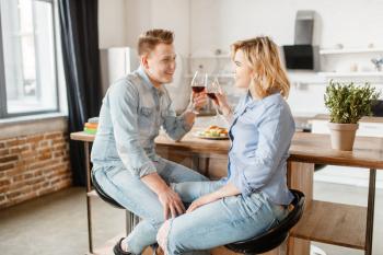 Attractive love couple sitting at the table, romantic dinner. Man and woman drinks wine and eats fruits on the kitchen. Happy lifestyle, beautiful relationship