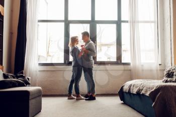 Young love couple with glasses of red wine standing at the window full of sunlight, romantic dinner. Man and woman relaxing in living room. Happy lifestyle, beautiful relationship