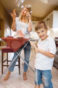 Housewife talking by phone, kid fooling with iron on the kitchen. Woman with child playing at home together. Female person with her playful son in their house