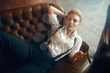 Woman in strict clothes lying on leather couch and smoking cigar, retro fashion, gangster style, female macho. Vintage business lady in office with brick walls