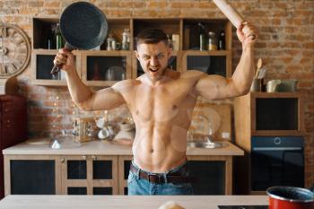 Muscular man with naked body holds frying pan and rolling pin on the kitchen. Nude male person preparing breakfast at home, food preparation without clothes