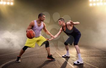Two basketball players playing the game on court, dark background with spotlights. Male athletes in sportswear play the game on streetball training