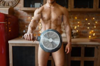 Nude man covered with a frying pan cooking romantic dinner on the kitchen. Naked male person preparing breakfast at home, food preparation without clothes