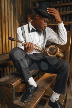 Black jazz musician sitting with saxophone, wooden fench background. Black jazzman in hat poses with instrument