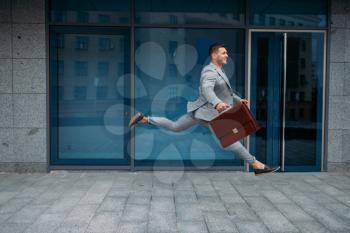 Happy businessman with briefcase jumping at the office building in downtown. Lucky business person in suit, urban style