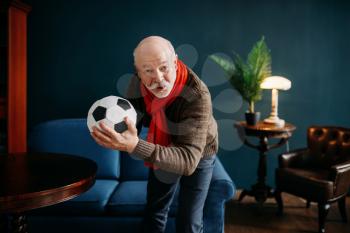 Elderly man with red scarf and ball watching TV, football fan. Bearded mature senior poses in living room, old age people leisure