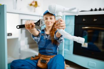 Young female plumber in uniform shows wrench and pipe in the kitchen. Handywoman with toolbag repair sink, sanitary equipment service at home