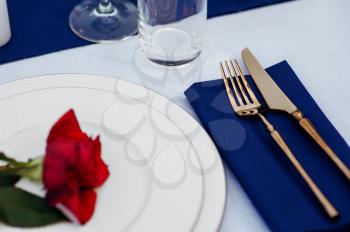 Table setting, silverware and red rose on the plate closeup, top view, nobody. Luxury banquet decoration, white tablecloth, tableware outdoors