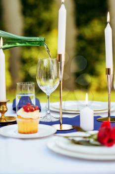 Table setting, wine is poured from a bottle into a wineglass, candles and flower on the plate closeup, nobody. Luxury silverware and white tablecloth, tableware outdoors