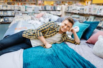 Young man lying on the bed in bedding store. Male person buying home goods in market, guy sleeping in shop