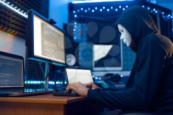 Hacker in mask and hood sitting at his workplace with laptop and PC, password or account hacking. Internet spy, illegal lifestyle, risk job, network criminal