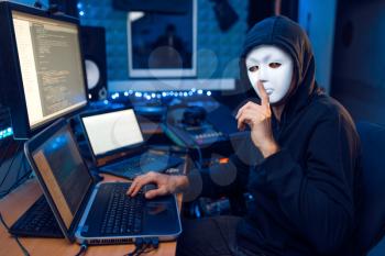 Hacker in mask and hood sitting at his workplace with laptop and PC, network or account hacking. Internet spy, illegal lifestyle, risk job