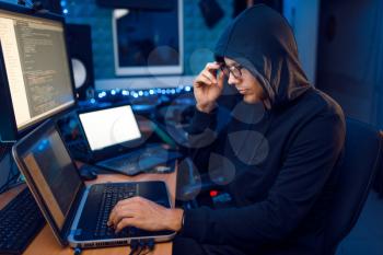 Hacker in hood at his workplace with laptop and desktop PC, website or corporate hacking, darknet user. Internet spy, crime lifestyle, risk job, network criminal