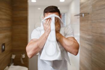 Man wipes his face with a towel, front view, routine morning hygiene. Male person at the sink performs skin and body treatment procedures