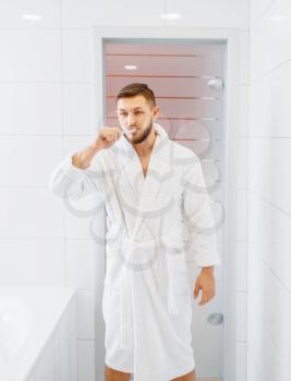 Man in bathrobe brushes his teeth in bathroom, routine morning hygiene. Male person at the sink performs skin and body treatment procedures